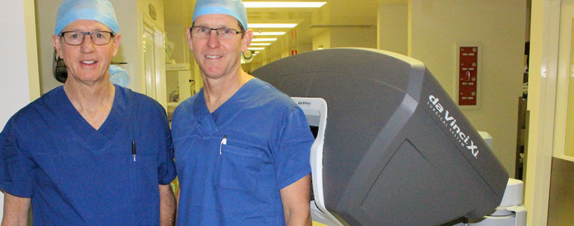 Buderim Private Hospital surgeons Dr John Hansen and Dr David Colledge wear blue scrubs as they stand beside the da Vinci robot