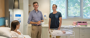 A Buderim Private Hospital doctor chats to a new mum lying in a hospital bed, and her partner, as their baby sleeps in a crib beside the bed.