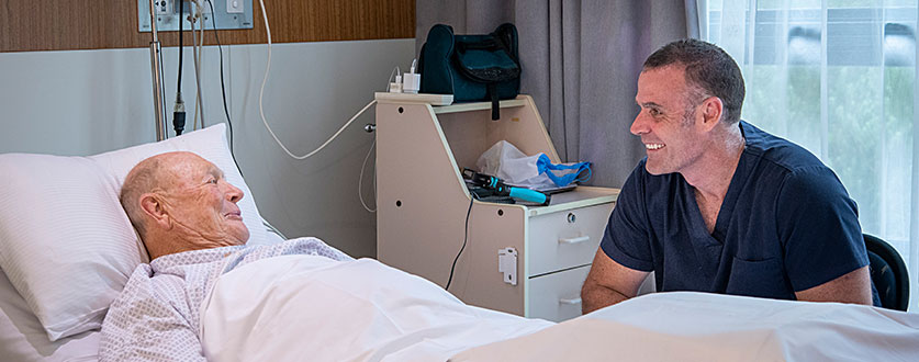 A male staff member chats with a male patient lying in bed at Buderim Private Hospital.